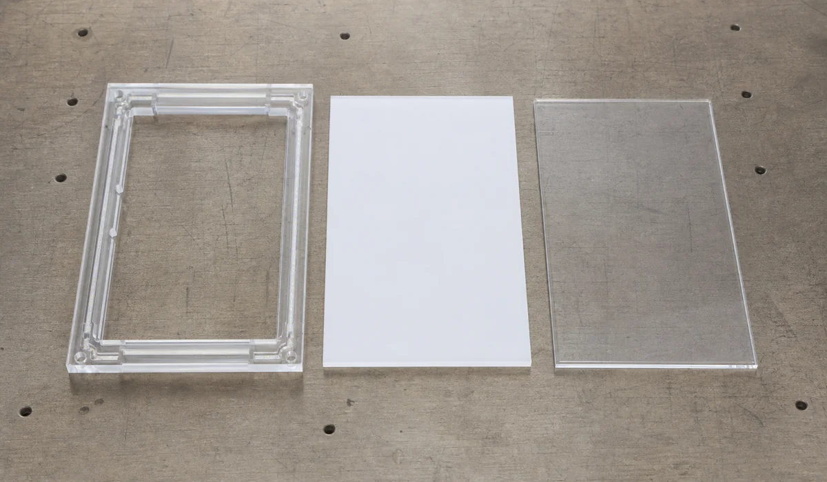 Flat-lay of a three-part acrylic light box frame kit on a workbench — CNC-machined frame body with visible LED channel, opal white diffuser sheet, and clear snap-off face panel