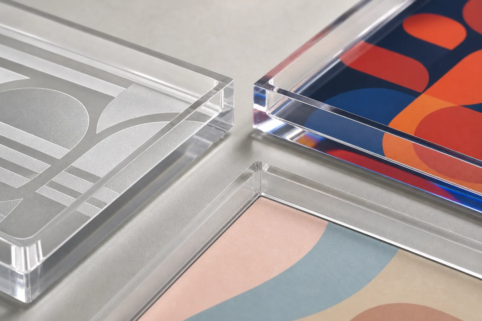 Corner details of three branded acrylic serving trays comparing acrylic tray logo options — frosted laser-etched monogram, vivid bottom-printed pattern under clear PMMA, and a replaceable printed insert edge visible in the tray base